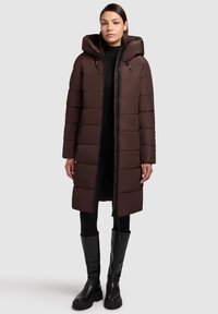 Doudoune marron avec une capuche, présentant un design matelassé, une fermeture éclair sur le devant et des poches latérales. Le modèle porte des leggings noirs et des bottes jusqu'aux genoux.