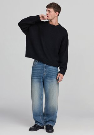 Giovane uomo che indossa un maglione nero oversize, jeans blu sbiaditi e larghi, e scarpe nere, in posa con una mano vicino alla bocca, guardando di lato.