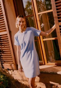 Νεαρή γυναίκα με ανοιχτό μπλε φόρεμα κοντών μανικιών στηρίζεται στο ξύλινο πλαίσιο παραθύρου μιας ρουστίκ πέτρινης κατοικίας σε ζεστό ηλιακό φως.