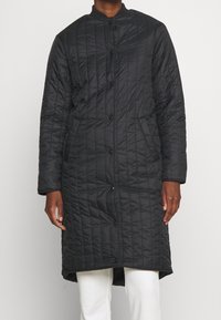 Abrigo largo negro acolchado con cuello alto, botones de presión en la parte delantera y tejido texturizado. Cuenta con mangas largas y una abertura lateral para facilitar el movimiento.