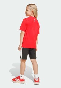 Červené tričko s krátkym rukávom a bielym logom, skombinované s čiernymi kraťasmi s bielymi detailmi, červenými teniskami a bielymi ponožkami s logom.