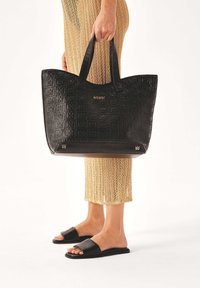 Melna ādas tote soma ar reljefa rakstu un zelta logo. Divas izturīgas rokturi un strukturēta forma. Valda kopā ar zelta adītu svārku.