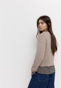 Beige kötött pulóver hosszú ujjakkal és kerek nyakkal, alatta sötét csíkos hosszú ujjú inggel párosítva. A modell laza kék farmert visel.
