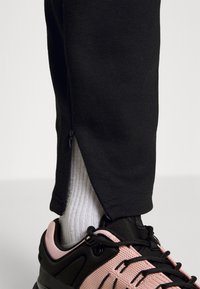 Svarta träningsbyxor med dragkedja i bensluten, tillsammans med svarta och rosa sneakers och vita räfflade strumpor. Närbild från ankeln.