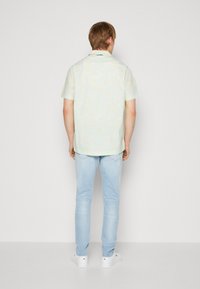 Camisa de manga corta con cuello en verde claro con un patrón sutil. Combinada con jeans azul claro y zapatillas blancas. De pie, visto desde la espalda.