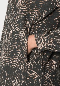 Schwarzer und beiger Stoff mit strukturiertem, tierischem Druckdesign. Nahaufnahme einer Hand, die in einer Seitentasche des Kleidungsstücks steckt.