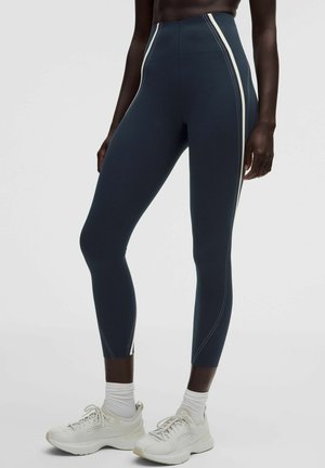 Person iført mørkeblå højttaljede leggings med hvide sidelister og hvide løbesko med sokker, stående på en lys baggrund.