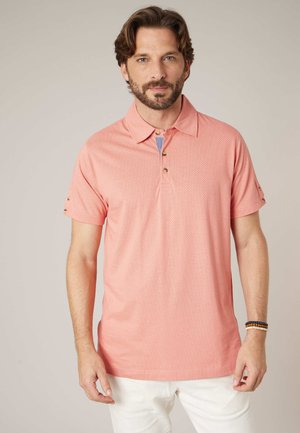 Homme aux cheveux bruns et à la barbe, portant un polo à manches courtes rose saumon et un pantalon blanc, debout devant un fond uni.