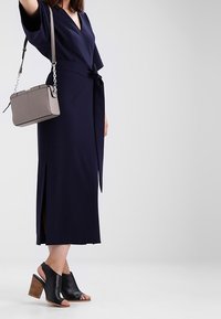 Námorně modré zavinovací šaty s krátkými rukávy a bočními rozparky, doplněné černými sandály s otevřenou špičkou a malou šedou crossbody taškou s řetízkem.