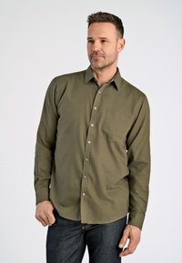 Olijfgroen overhemd met lange mouwen, voorzien van een knoopsluiting, een borstzak en grijze knopen, gecombineerd met donkerdenim jeans.