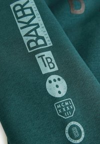 Tume teal kangas prinditud graafikaga: "BAKER," "TB," ja aasta 1983 erinevates fontides, lisaks ringiline kujundus punktidega. Tekstuuriga pind.