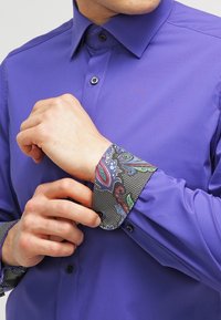 Paarse overhemd met een gladde textuur, voorzien van een zwarte manchet met paisleypatroon. De afgebroken kraag en knoopdetails zijn zichtbaar.