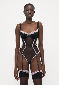 Lingerie nera in rete a pois trasparente, con dettagli in pizzo rosa, spalline arricciate e gancetti per giarrettiere regolabili. Coppe con ferretto per un buon supporto.