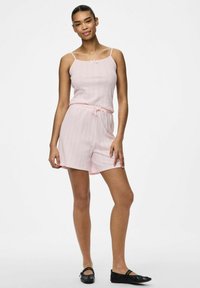 Mono sin mangas color rosa claro con una tela texturizada, un acento de lazo en el escote y una cintura con cordón. Unos zapatos planos negros completan el conjunto.