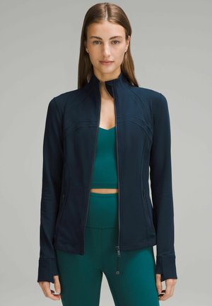 Marineblå zip-up jakke med høj krave, tætsiddende design og lange ærmer. Blødt, strækbart materiale med sømdetaljer og tommelfingerhuller ved manchetterne.