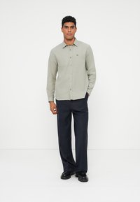Un uomo è in piedi contro uno sfondo bianco, indossa una camicia grigio chiaro con bottoni, pantaloni larghi neri e scarpe nere, con una mano in tasca.