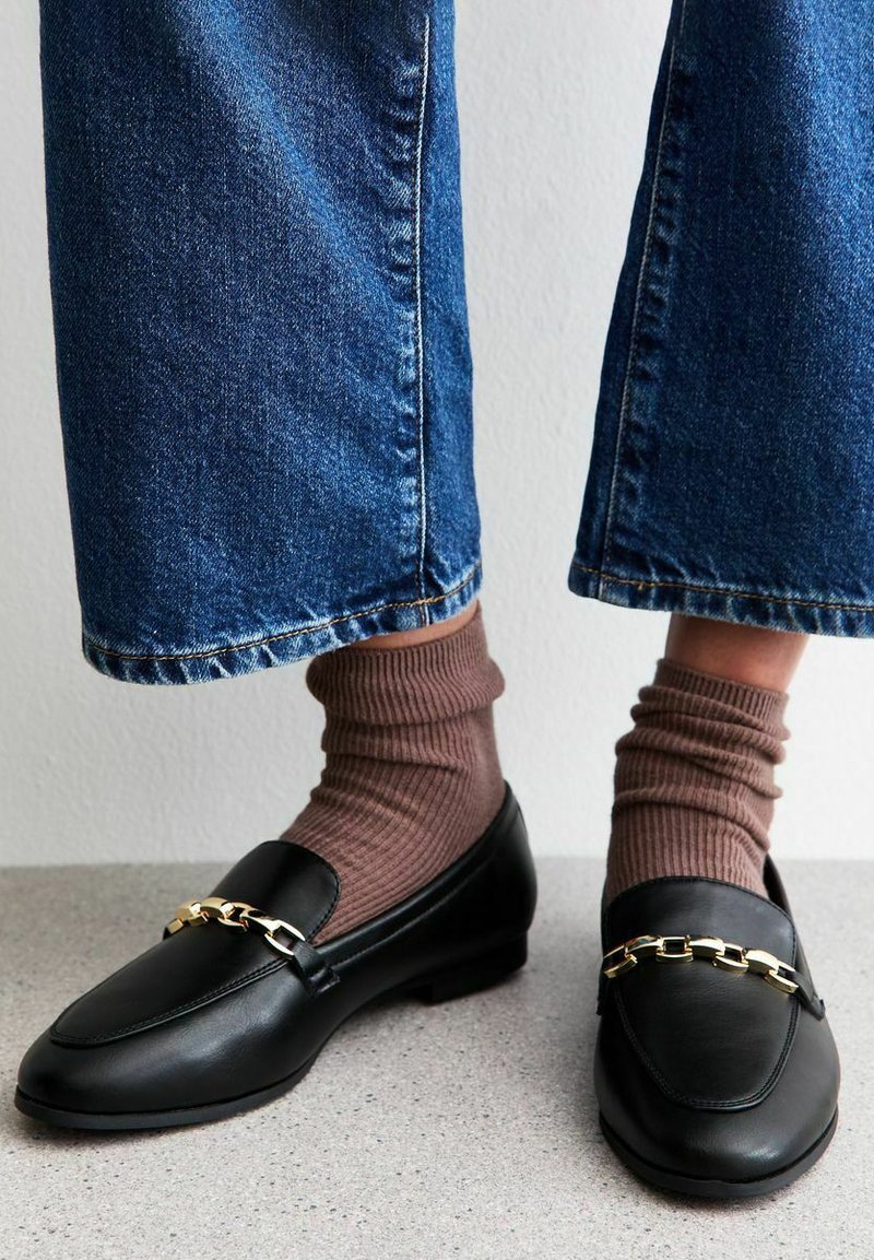 Mocasines de cuero negro con un acento de cadena dorada, usados con calcetas marrones acanaladas, combinados con jeans de mezclilla azul de corte acampanado. Acabado texturizado y suave.