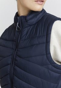 Tmavě modrá vatovaná vesta s vysokým límcem, diagonálním prošívaným vzorem a předním zipem. Texturovaný, lehký materiál.