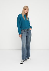 Sudadera color azul verdoso con un corte relajado y puños acanalados, combinada con unos jeans de tiro alto y pierna ancha de color azul y zapatillas en blanco y negro.