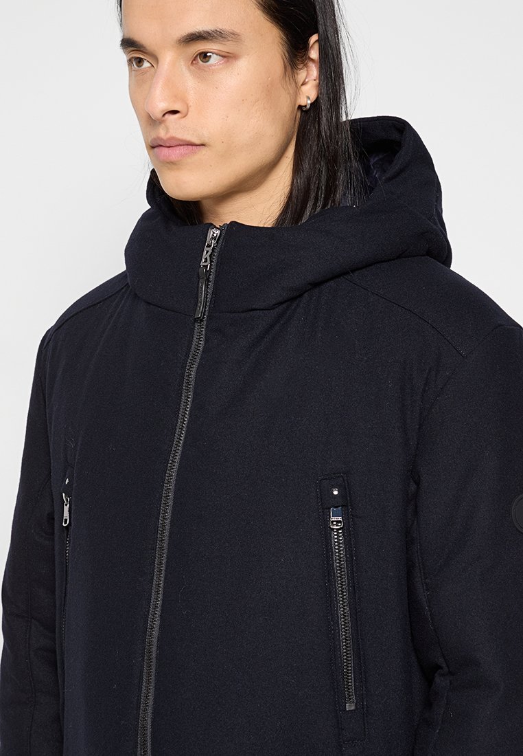 Schwarze Kapuzenjacke mit strukturiertem Finish, ausgestattet mit Reißverschluss-Taschen und einem hohen Kragen für Wärme.