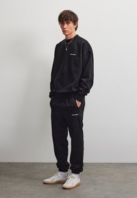 Joven vestido con sudadera negra Pegador y pantalones deportivos, con zapatillas blancas, de pie contra pared gris y suelo de hormigón.
