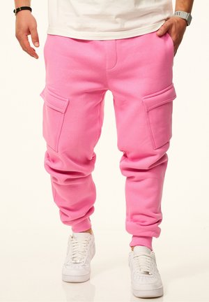 Mann trägt knallpinke Cargo-Sweatpants mit großen Seitentaschen und weißen Turnschuhen, Hände entspannt an den Seiten.