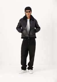 Schwarze Lederbomberjacke mit Schafwollkragen, Reißverschluss und seitlichen Taschen, kombiniert mit locker sitzenden schwarzen Jeans und weißen Sneakers.