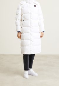 Vit puffjacka med lång design, utrustad med horisontell quiltning, sidofickor och huva. Bärs över mörka jeans och vita sneakers.