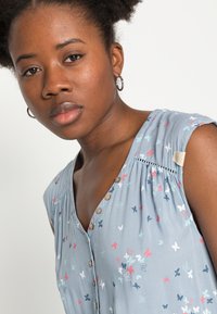 Blusa sem mangas azul com padrões de borboleta em vermelho, branco e azul. Apresenta um fecho frontal com botões e detalhes franzidos na gola.
