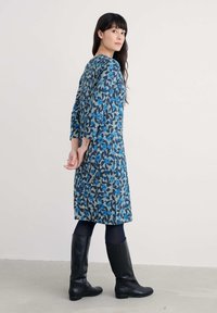 Rochie cu imprimeu floral albastru, cu manșete trei sferturi, până la genunchi, dintr-un material moale. Purtată cu cizme negre până la genunchi.