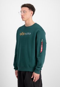 Tamnozeleni dukserica od pamuka s rebrastim okruglim izrezom, koja ima zlatni logo "Alpha Industries" i džep sa rajsferšlusom na rukavu s crvenom oznakom.