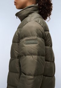 Olijfgroen pufferjack met een hoge kraag, voorzien van gevoerde secties, ritssluiting zijzakken en een gestructureerde logo label op de mouw.