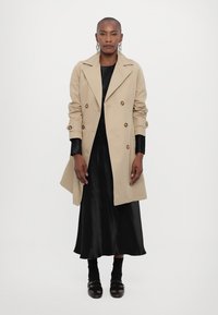 Beige trench coat with double-breasted buttons, wide lapels, and adjustable cuffs, layered over a black satin dress and black shoes.