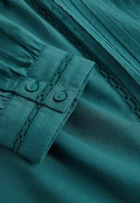 Tissu bleu sarcelle avec un poignet boutonné et un détail de dentelle, mettant en valeur la texture et les coutures d'une manche de vêtement.