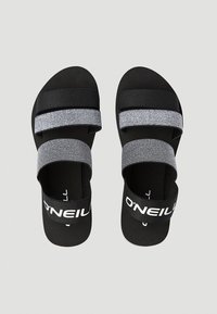 Mustat sandaalit, joissa on kaksi teksturoitua harmaata remmiä ja musta brändiremmi. Ominaisuuksiin kuuluu litteä pohja ja minimalistinen muotoilu.