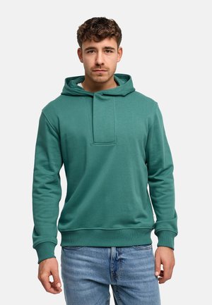 Jeune homme aux cheveux courts et bouclés portant un sweat à capuche bleu sarcelle et un jean bleu clair, debout devant un fond blanc uni.