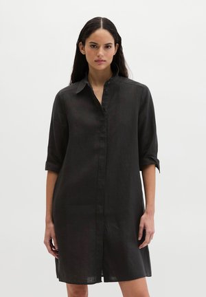 Frau mit langen dunklen Haaren trägt ein schwarzes Hemdkleid mit Dreiviertelärmeln vor einem schlichten hellen Hintergrund.