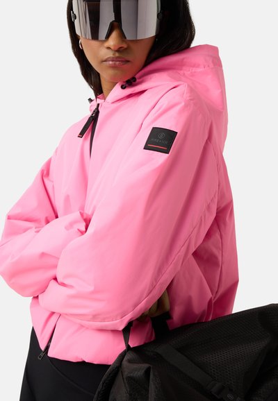 Person trägt übergroße reflektierende Sonnenbrille und eine leuchtend pinke Kapuzenjacke mit einem schwarzen Fleck am Ärmel, hält eine schwarze strukturierte Tasche.