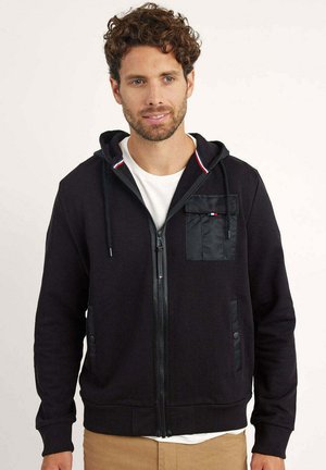 Homme aux cheveux bouclés portant un sweat à capuche noir zippé avec poches avant et un t-shirt blanc, debout devant un fond uni.