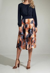 Femme portant un haut noir à manches longues associé à une jupe jusqu'aux genoux présentant un motif géométrique abstrait marron, beige et bleu, ainsi que des talons marron.