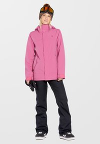 Roze sneeuwjack met een aansluitend ontwerp, voorzien van twee zijzakken, gecombineerd met zwarte sneeuwbroeken en zwarte sportieve schoenen. Een gebreide muts en een skibril maken de outfit compleet.