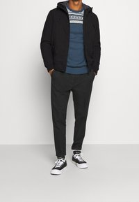 Černá bunda s kapucí, modrý svetr s bílým fair isle vzorem, šedé kalhoty a černé tenisky s bílou podrážkou.