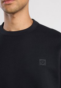 Felpa nera con scollo tondo, tessuto texturizzato e un piccolo patch quadrato con un logo a forma di diamante nella zona del petto sinistro.