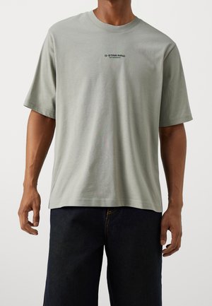 Mand iført lysegrå G-Star Raw kortærmet T-shirt og mørkeblå jeans, stående med arme afslappet ned langs siderne.