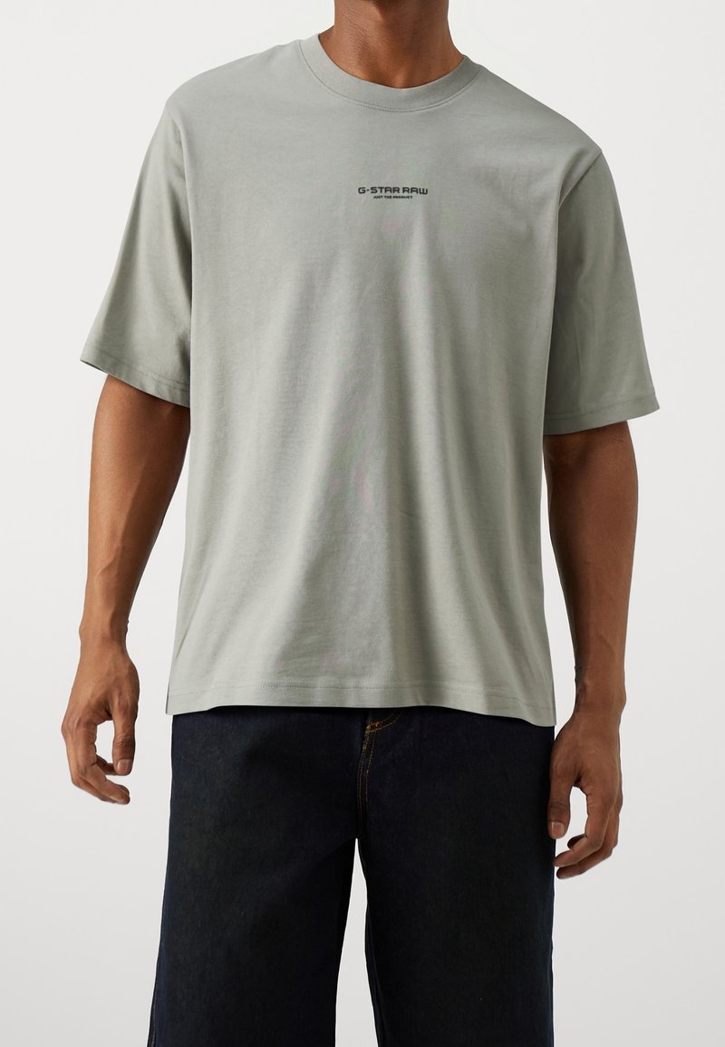 Homme portant un t-shirt à manches courtes G-Star Raw gris clair et un jean bleu foncé, debout, les bras détendus le long du corps.