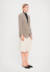 Beige skreddersydd blazer med en enkelt knapp, kombinert med en svart turtleneck, kremfargede, plissert shorts og svarte kitten-heel mules.