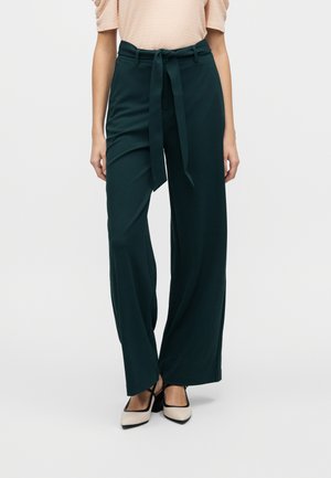Bukser med vide ben i mørkegrønn, med stoffbelte, glatt tekstur og løs passform. Kombinert med lyse, blokkhælte sko.