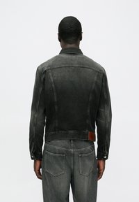 Mørkegrå denimjakke med tætsiddende pasform, tydelige sømlinjer og et brunt lædermærke. Har knaplukning ved ærmekanterne og to baglommer.