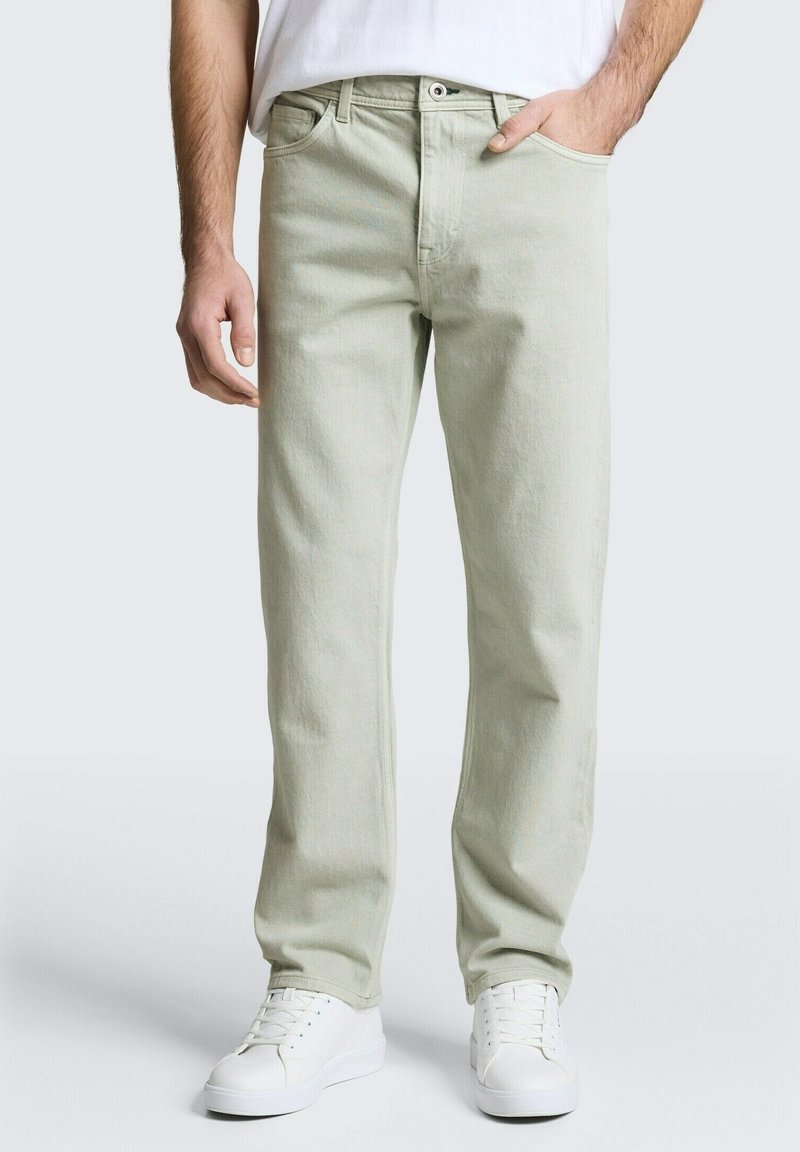 Homme portant un jean beige clair droit et des baskets blanches, debout avec une main dans la poche, sur fond uni.