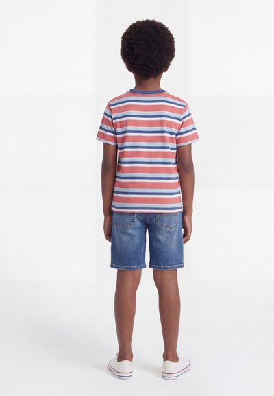 Αγόρι που στέκεται πλάτη, φοράει κοντομάνικη ριγέ μπλούζα, τζιν σορτς και λευκά αθλητικά παπούτσια σε απλό λευκό φόντο.